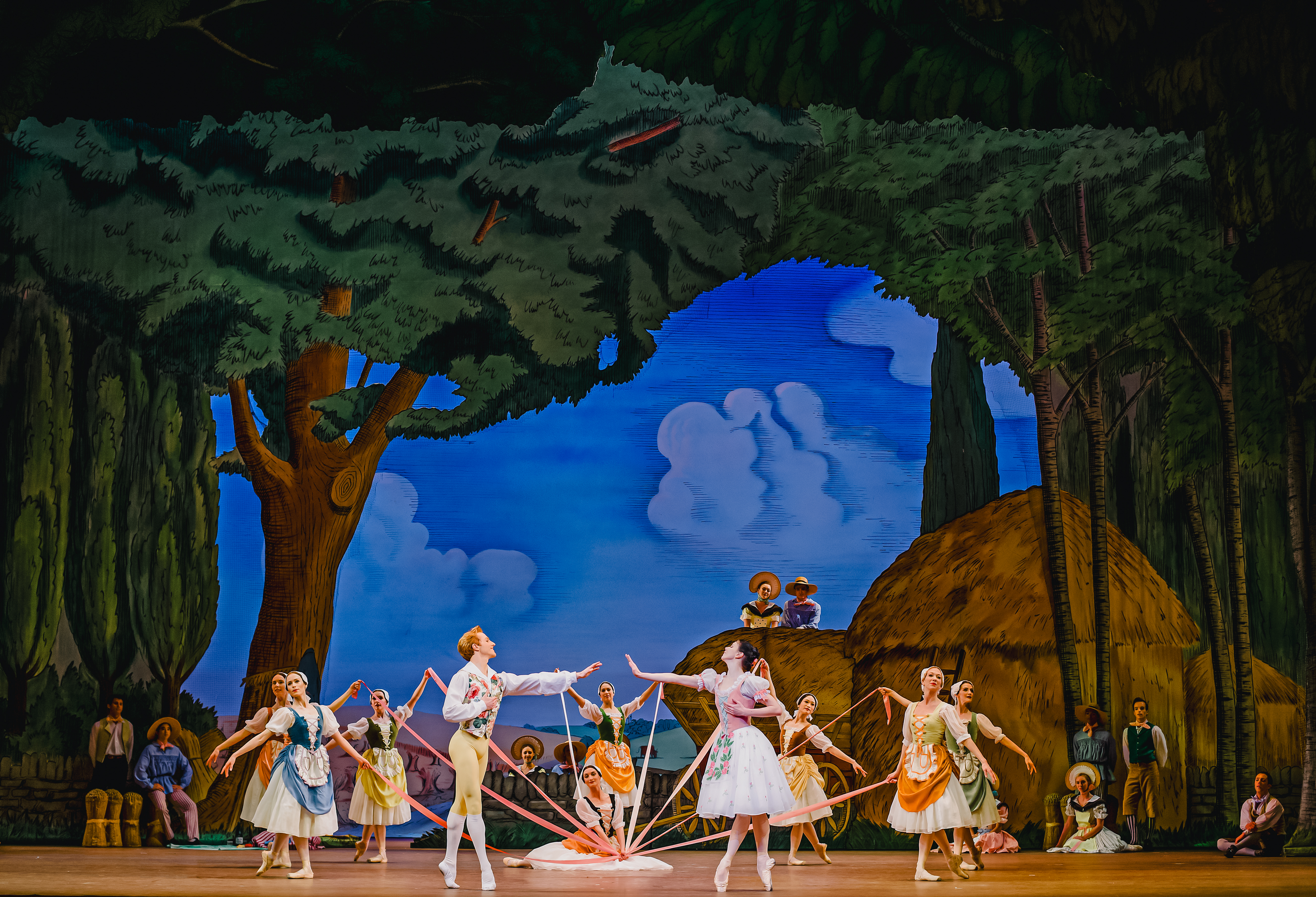 The stage of The Royal Opera House is set for a performance of La Fille mal gardée. The scenery looks as though it has been painted in an Impressionist style and shows trees in a forest, hay bales and a bright blue sky with blurred blue clouds. In the centre of the stage several dancers of The Royal Ballet stand around a maypole, other dancers are in the foreground observing. The dancers wear rural-style corset dresses and pointe shoes. At the front of the maypole 