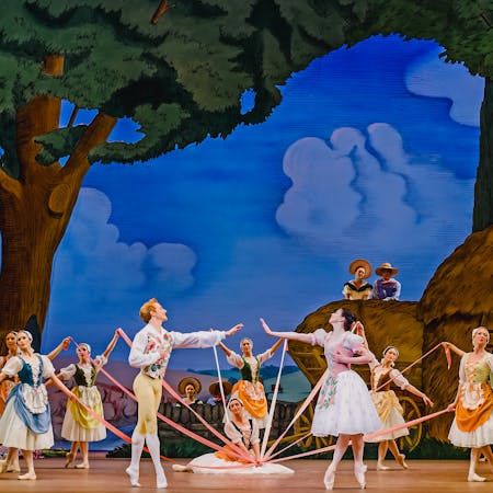 The stage of The Royal Opera House is set for a performance of La Fille mal gardée. The scenery looks as though it has been painted in an Impressionist style and shows trees in a forest, hay bales and a bright blue sky with blurred blue clouds. In the centre of the stage several dancers of The Royal Ballet stand around a maypole, other dancers are in the foreground observing. The dancers wear rural-style corset dresses and pointe shoes. At the front of the maypole