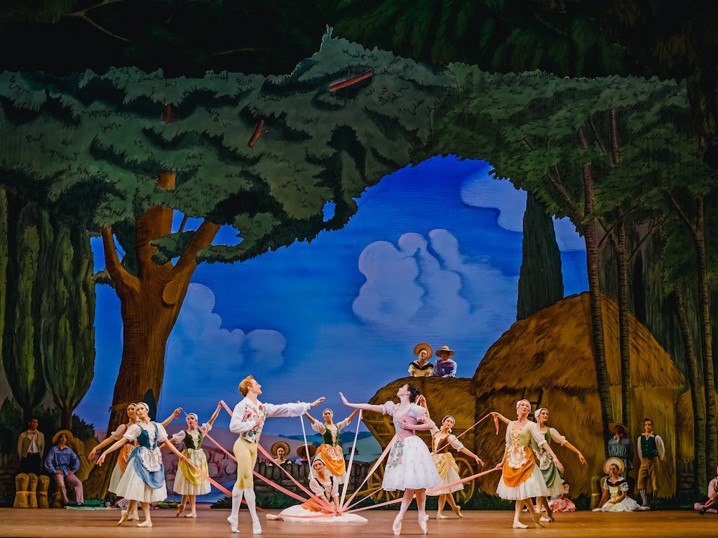 The stage of The Royal Opera House is set for a performance of La Fille mal gardée. The scenery looks as though it has been painted in an Impressionist style and shows trees in a forest, hay bales and a bright blue sky with blurred blue clouds. In the centre of the stage several dancers of The Royal Ballet stand around a maypole, other dancers are in the foreground observing. The dancers wear rural-style corset dresses and pointe shoes. At the front of the maypole