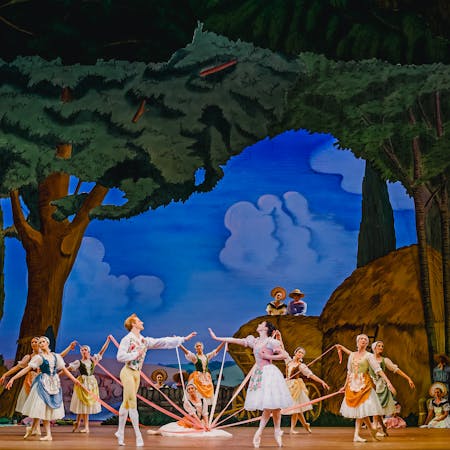 The stage of The Royal Opera House is set for a performance of La Fille mal gardée. The scenery looks as though it has been painted in an Impressionist style and shows trees in a forest, hay bales and a bright blue sky with blurred blue clouds. In the centre of the stage several dancers of The Royal Ballet stand around a maypole, other dancers are in the foreground observing. The dancers wear rural-style corset dresses and pointe shoes. At the front of the maypole