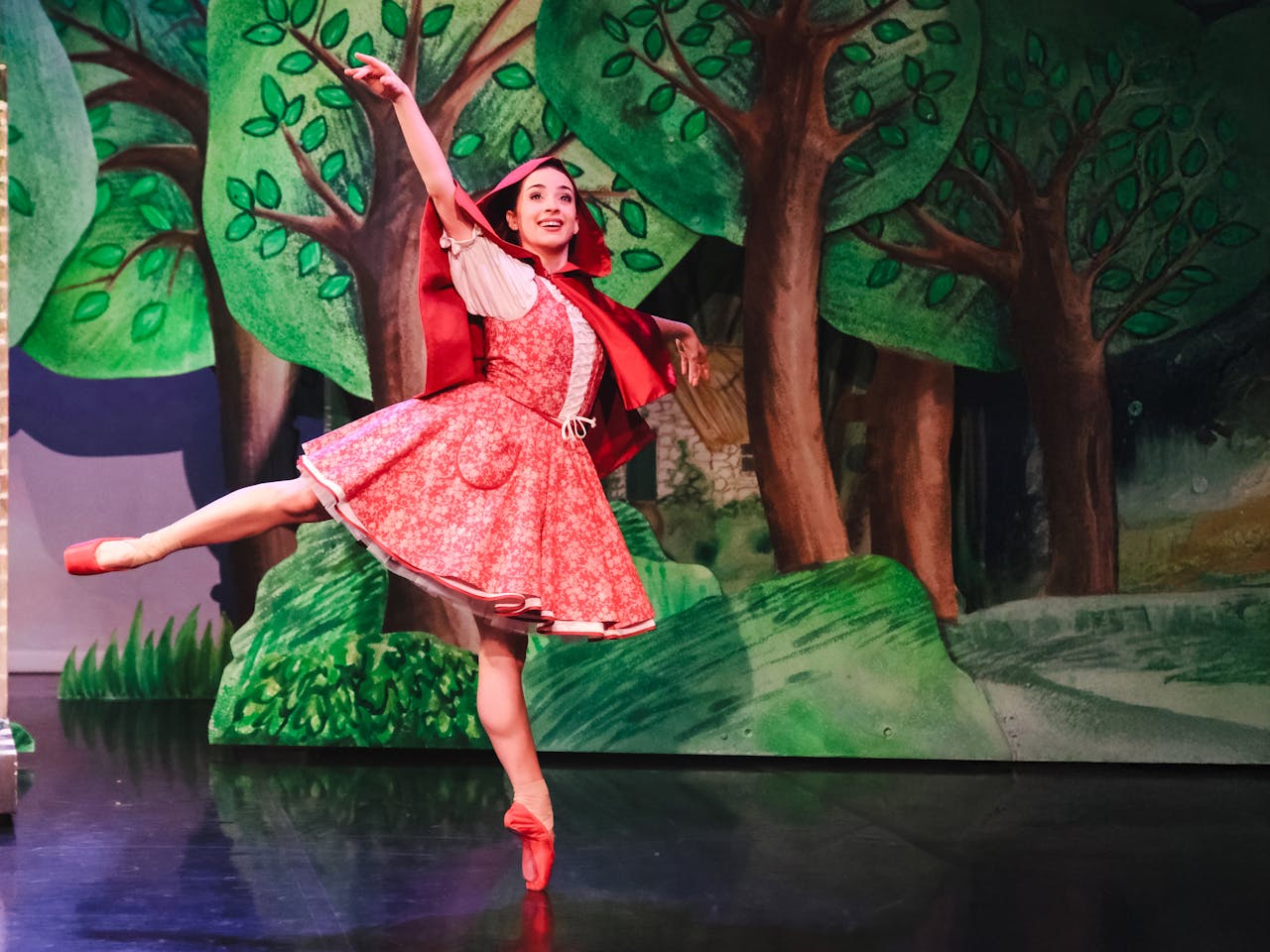 The ballet dancer Julie Nunès wears red pointe ballet slippers, a red and white flower-patterned corseted dress and a red cape. She stands en pointe on her left leg with her right leg pointing behind her as she performs in The Royal Ballet and Northern Ballet’s Little Red Riding Hood. The stage is set with tall hand drawn green trees that overlap one another so they appear 3D.
