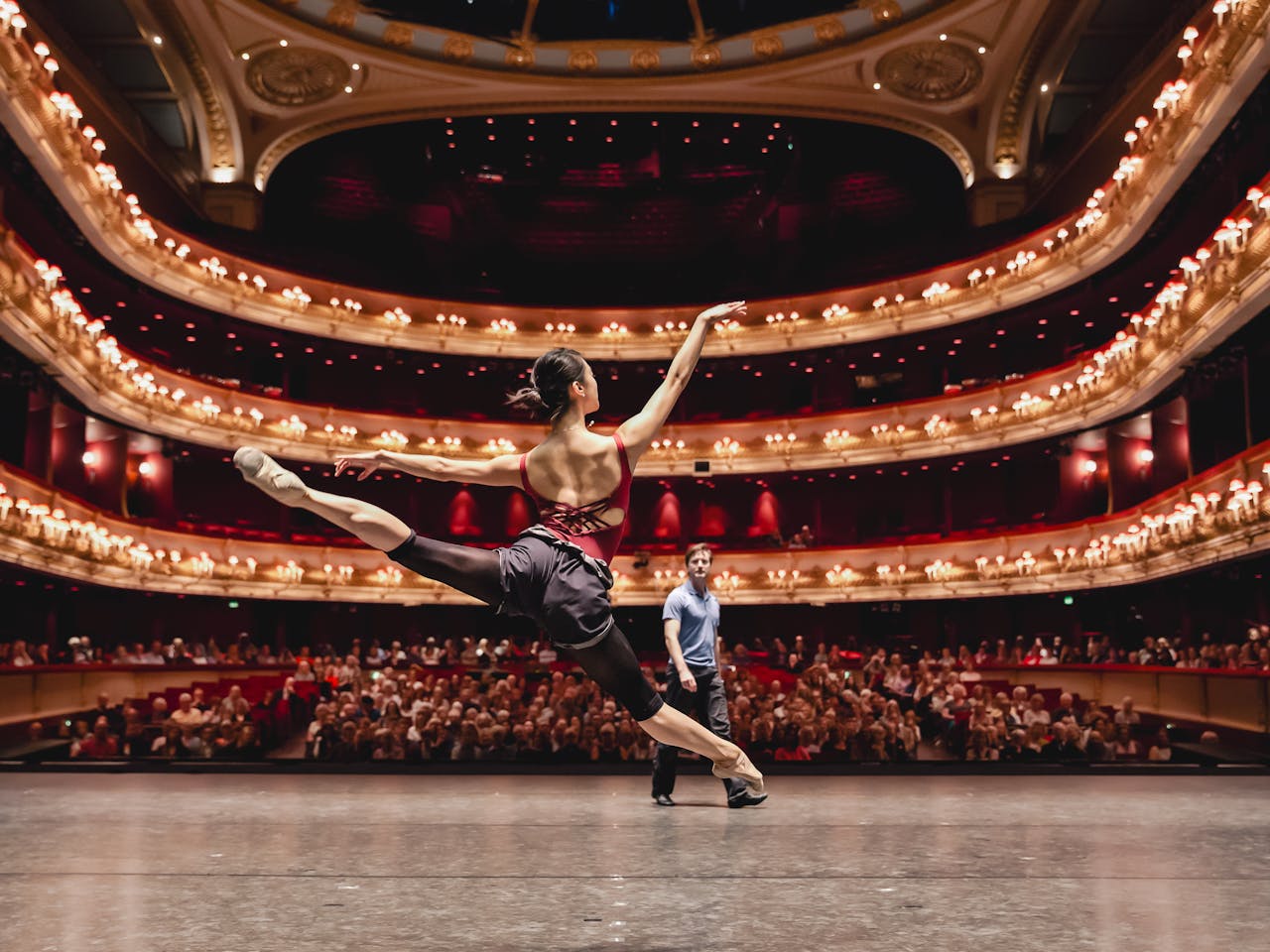 The Royal Ballet Dancer Fumi Kaneko performs in the centre of the stage of The Royal Opera House to an audience that are visible from the back of the stage. She launches herself into the air with her right leg straight ahead and her toes en pointe and her left leg behind her to create a line. Her right arm is stretched outwards above her shoulders and her left arm stretched out behind to follow the same line shape as her legs. In front of her stands a