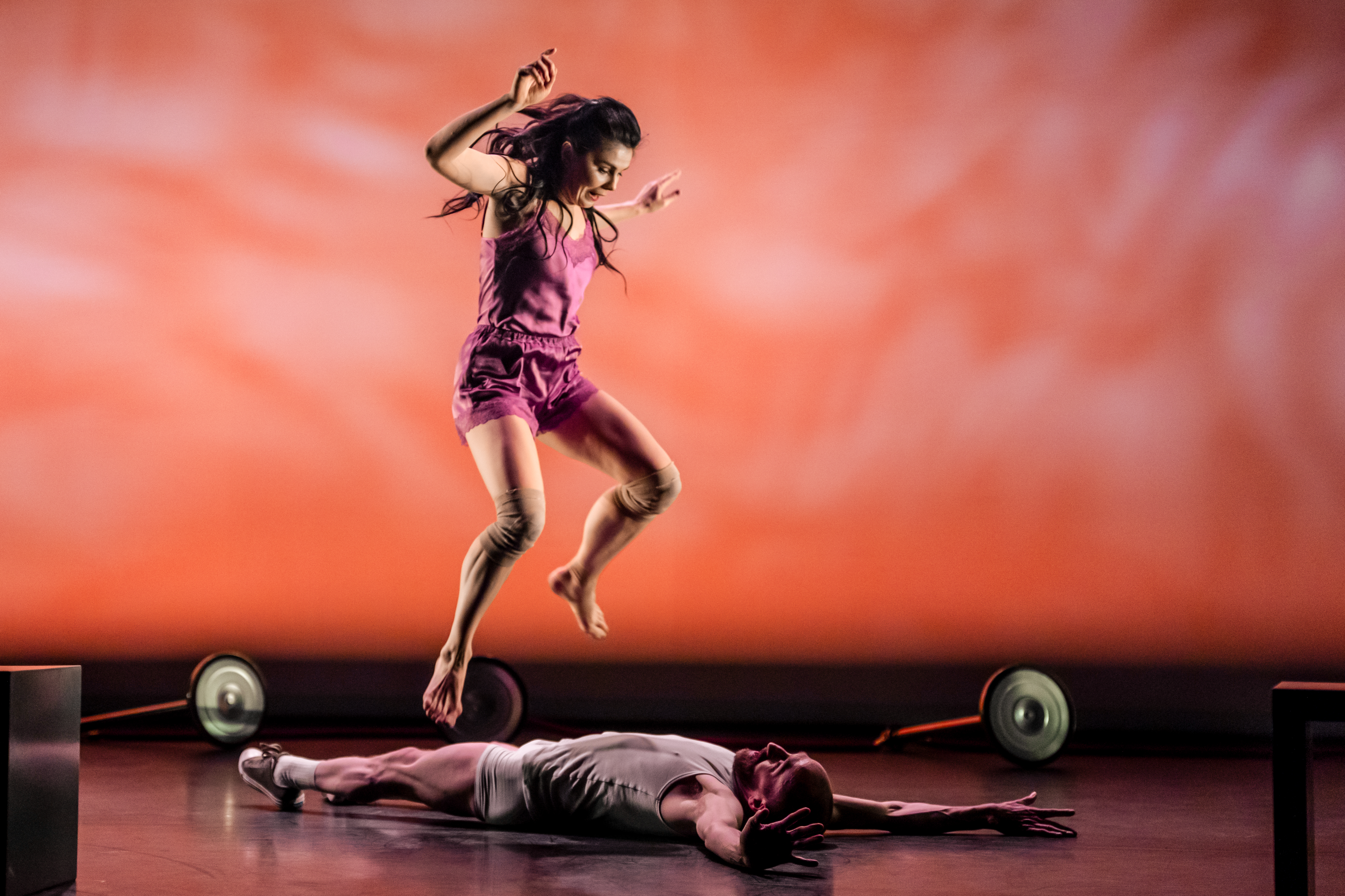 The ballet dancer Natalia Osipova, wearing a pink top and shorts jumps over the dancer Christopher Akrill who is lying on the floor on his back facing upwards  with his arms stretched above his head and wearing a white vest, shorts and trainers. The stage is set with a painterly-effect orange background and there are two wheels scattered around them.
