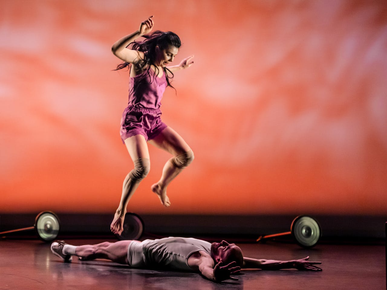 The ballet dancer Natalia Osipova, wearing a pink top and shorts jumps over the dancer Christopher Akrill who is lying on the floor on his back facing upwards with his arms stretched above his head and wearing a white vest, shorts and trainers. The stage is set with a painterly-effect orange background and there are two wheels scattered around them.