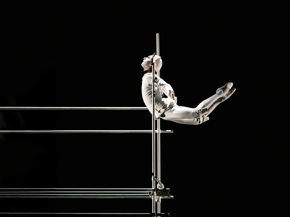A ballet dancer wearing all white with a white painted face to look like a clown holds onto scaffolding in a black-scened theatre. They create a C-shaped form with their body as their legs stretch out behind them.