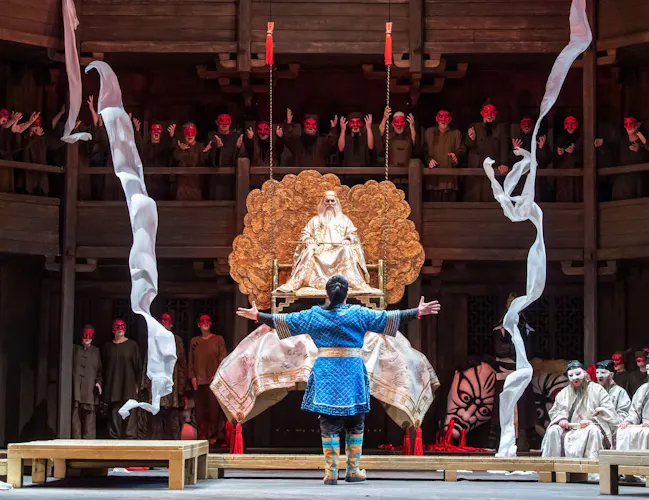 A performer with long white hair and wearing a long gold cloak holds a long baton on their lap. They are sitting on a gold chair on a raised on a platform with gold detailing behind. In front of them stands a performer wearing a blue jacket and patterned boots. They have their back to the audience and stand facing the person raised on the platform. Around them a chorus of performers wear a variety of masks and hold their hands out towards those in the centre.