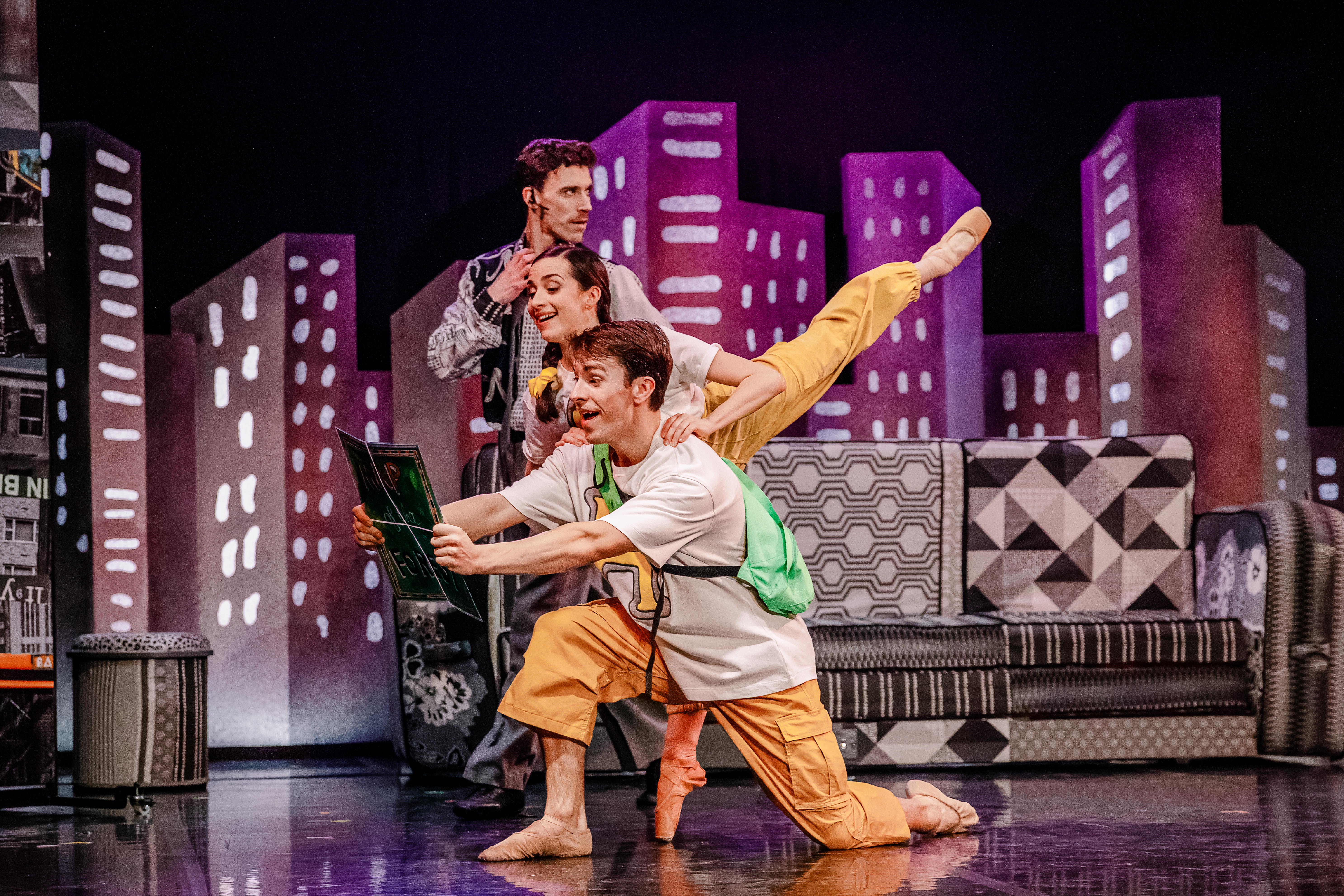 A theatrical performance of the ballet Hansel and Gretel by Northern Ballet depicts three performers on stage, Archie Sherman, Julie Nunès and Andrew Tomlinson. Behind them is a colorful backdrop of stylized city buildings. Archie Sherman is the performer at the front and is wearing a white t-shirt with a green backpack and orange pants. He kneels on one knee, holding a large book with a surprised expression. Julie Nunès is in a white top and yellow pants and stands behind him, balancing gracefully with one leg extended in an arabesque position. Andrew Tomlinson, dressed in a gray patterned suit, stands behind them with a headset microphone, looking on with a curious expression. The stage setting includes patterned furniture and vibrant, geometric scenery, evoking a whimsical cityscape.