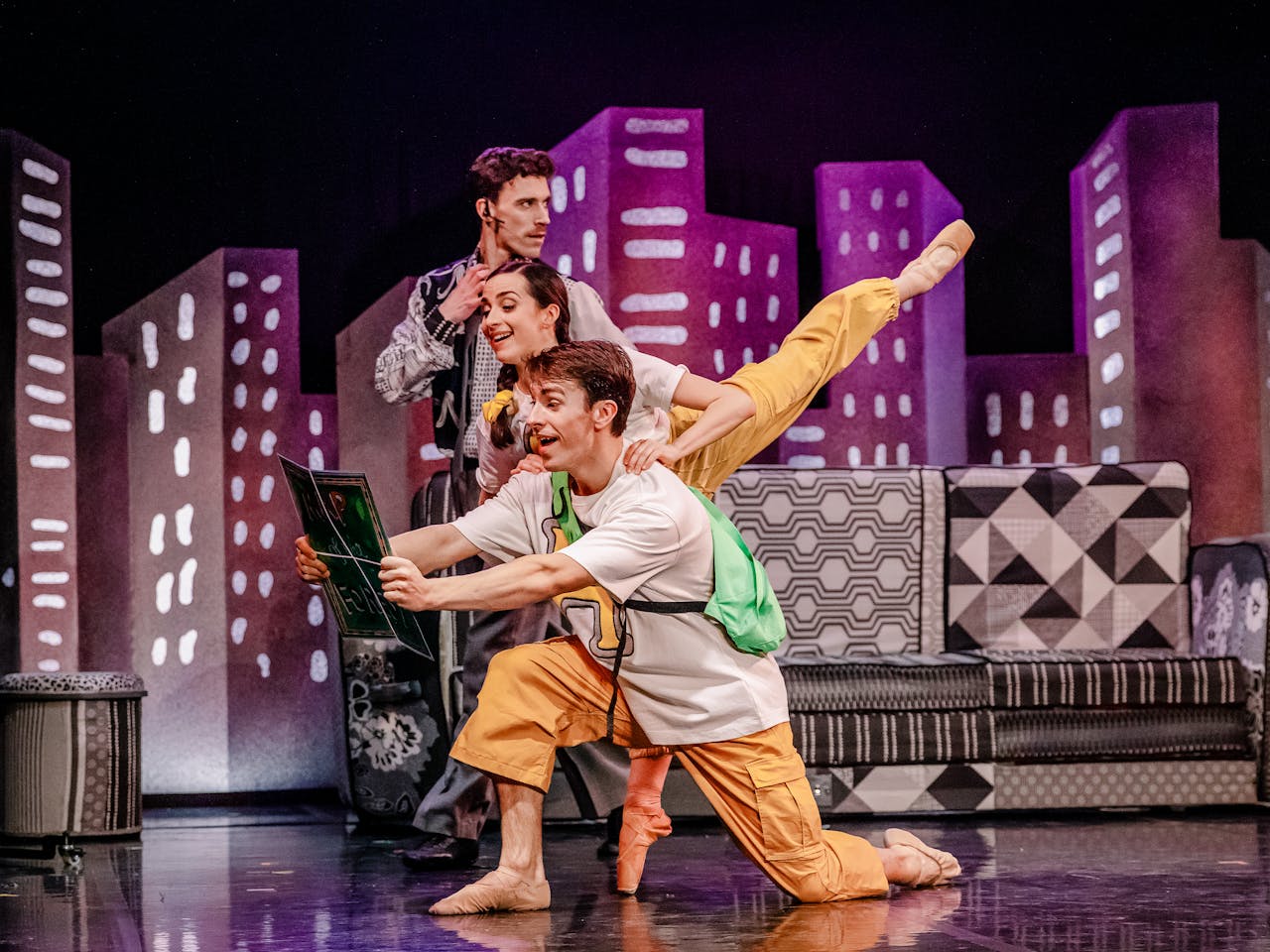 A theatrical performance of the ballet Hansel and Gretel by Northern Ballet depicts three performers on stage, Archie Sherman, Julie Nunès and Andrew Tomlinson. Behind them is a colorful backdrop of stylized city buildings. Archie Sherman is the performer at the front and is wearing a white t-shirt with a green backpack and orange pants. He kneels on one knee, holding a large book with a surprised expression. Julie Nunès is in a white top and yellow pants and stands behind him, balancing gracefully with one leg extended in an arabesque position. Andrew Tomlinson, dressed in a gray patterned suit, stands behind them with a headset microphone, looking on with a curious expression. The stage setting includes patterned furniture and vibrant, geometric scenery, evoking a whimsical cityscape.
