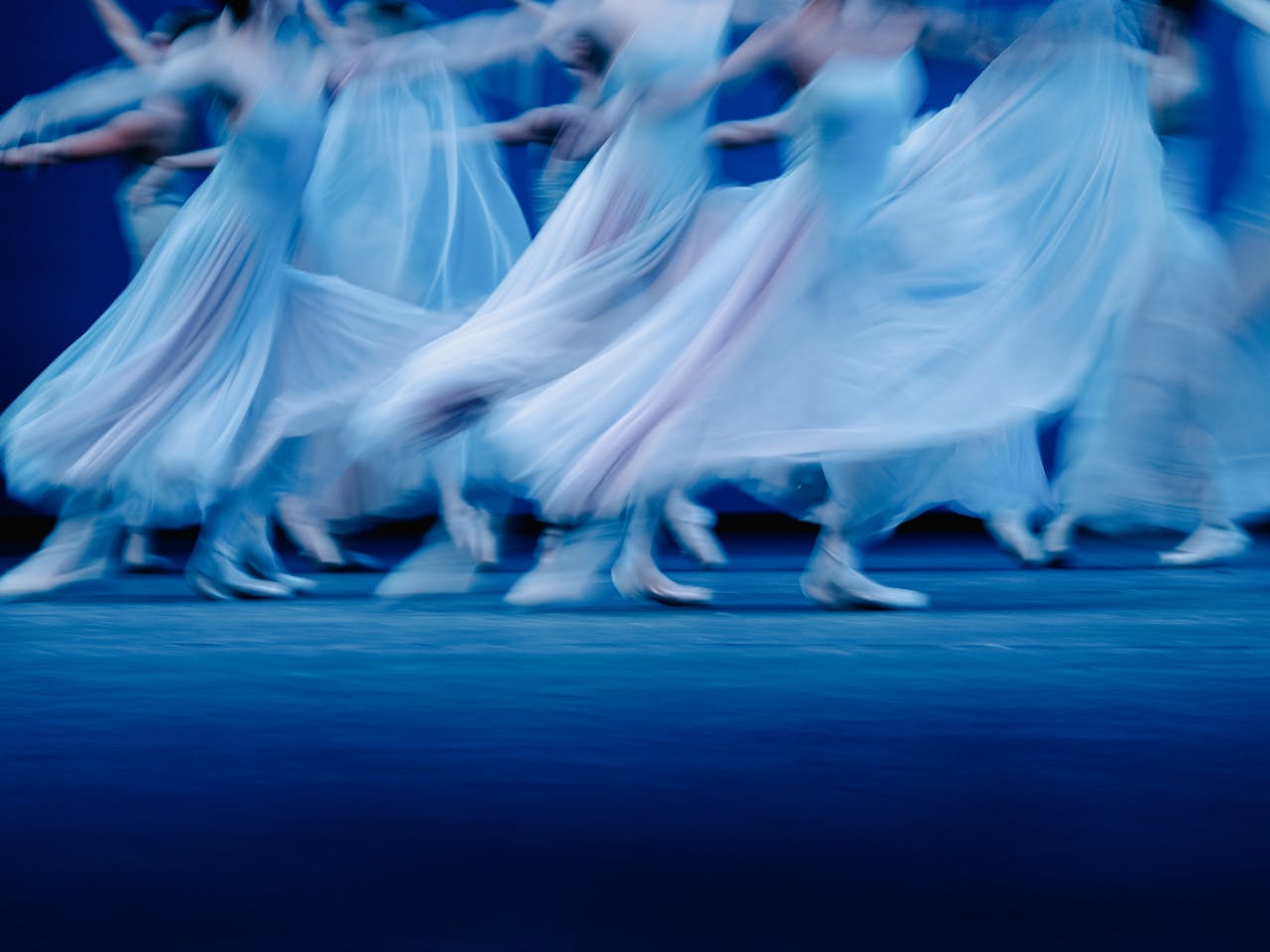 Blurred motion photograph of dancers of The Royal Ballet in flowing white dresses performing Balanchine’s Serenade on a dimly lit stage. The dancers' movements create a dreamy and ethereal effect, with their skirts billowing as they glide across the floor. The soft blue lighting enhances the sense of grace and fluidity in the performance.