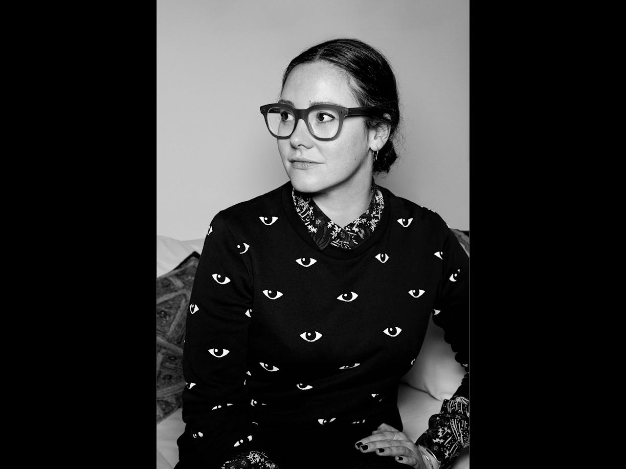 A black and white headshot of the set designer Chloe Lamford. She sits on a sofa and wears thick black rimmed glasses, a black top with eyes printed all over and a flower-patterned shirt peeks out from the jumper.
