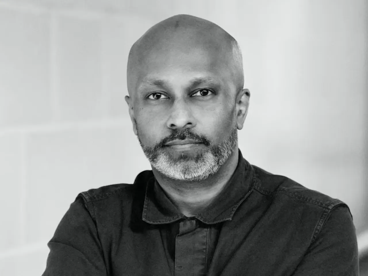 A black and white headshot of Akram Khan. He is bald and has a beard. He is wearing a black shirt.