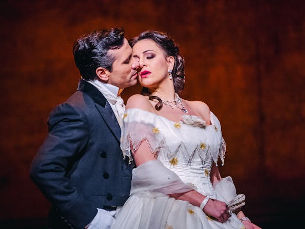 The opera singer Joyce El-Khoury performs as Violetta Valéry in The Royal Opera’s production of La traviata. She wears a whitw off shoulder ballgown with yellow embroidered flowers at the bust, a diamond antique-style necklace and she have a white chiffon scarf draped over her forearms. In her hand she holds a fan. She leans against the opera singer Sergey Romanovsky who is performing as Alfredo Germont who is wearing a navy-blue tailed coat and white shirt. His head is facing Violetta passionately, as though they are tenderly embracing.