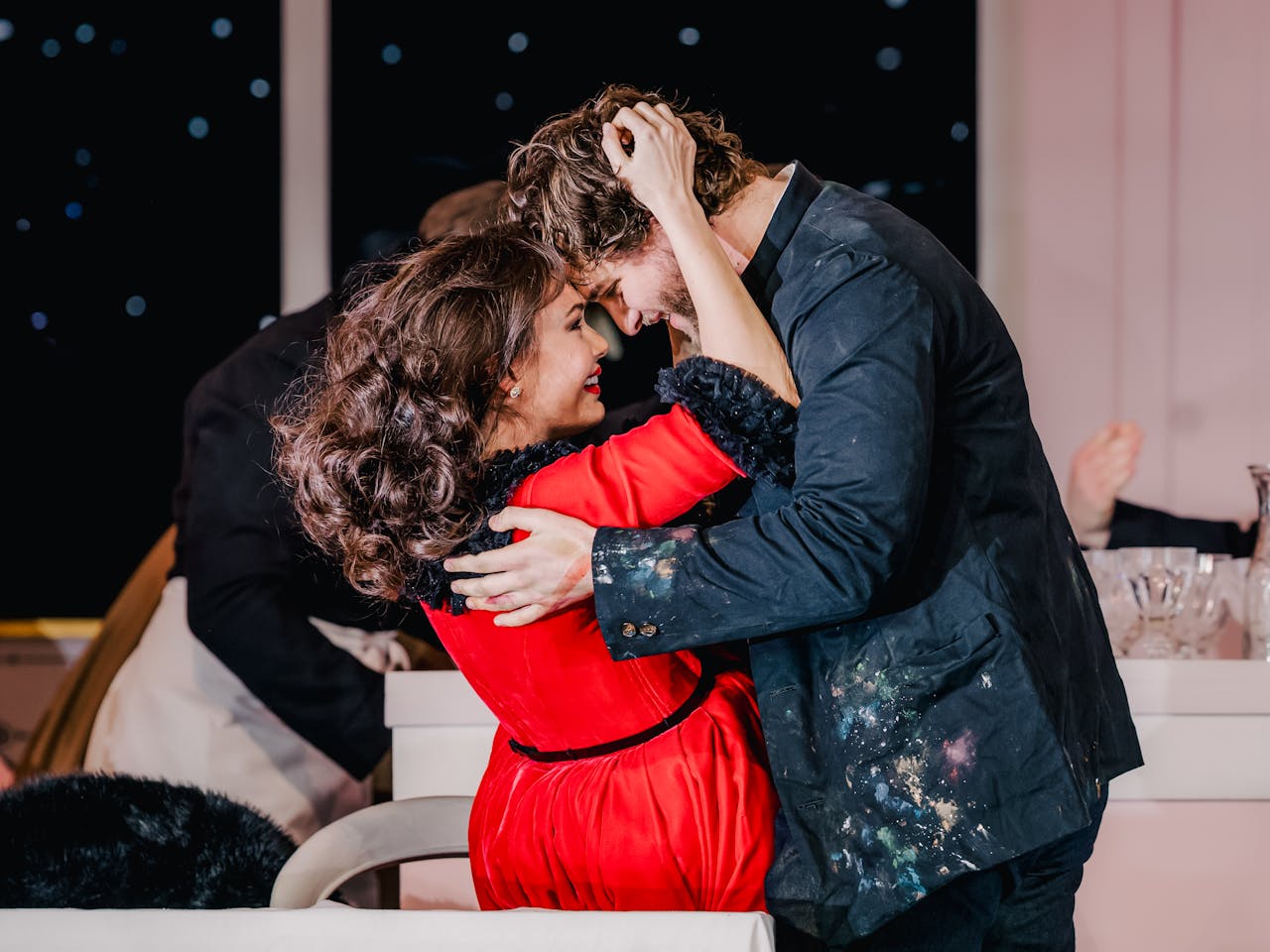 Two performers romantically embrace. One has long brown hair and is wearing a red dress with black fur lining and she holds onto the head of the other performer who is wearing a paint-specked artist smock jacket. They are Aida Garifullina performing as Musetta and Andrzej Filończyk as Marcello in The Royal Opera’s La bohème.
