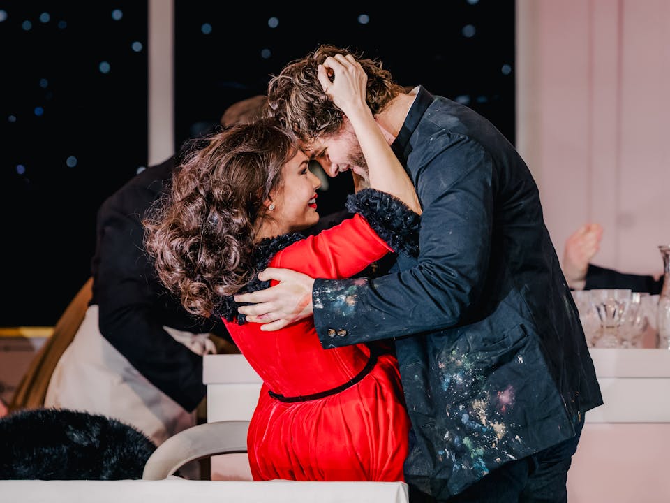 Two performers romantically embrace. One has long brown hair and is wearing a red dress with black fur lining and she holds onto the head of the other performer who is wearing a paint-specked artist smock jacket. They are Aida Garifullina performing as Musetta and Andrzej Filończyk as Marcello in The Royal Opera’s La bohème.