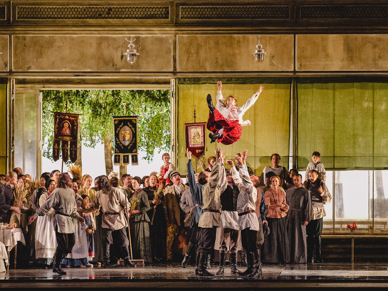 A lively and celebratory scene from The Metropolitan Opera’s production of Eugene Onegin. A group of men dressed in white rustic shirts and dark pants toss a young woman, dressed in a white blouse and red skirt, high into the air. The background features a large gathering of people in traditional folk attire, holding banners with religious imagery and symbols, and carrying wheat sheaves. The open doors reveal lush green trees outside, enhancing the festive atmosphere. The setting appears to depict a community celebration, possibly a wedding or harvest festival, with joyful expressions and dynamic movement.