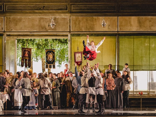 A lively and celebratory scene from The Metropolitan Opera’s production of Eugene Onegin. A group of men dressed in white rustic shirts and dark pants toss a young woman, dressed in a white blouse and red skirt, high into the air. The background features a large gathering of people in traditional folk attire, holding banners with religious imagery and symbols, and carrying wheat sheaves. The open doors reveal lush green trees outside, enhancing the festive atmosphere. The setting appears to depict a community celebration, possibly a wedding or harvest festival, with joyful expressions and dynamic movement.