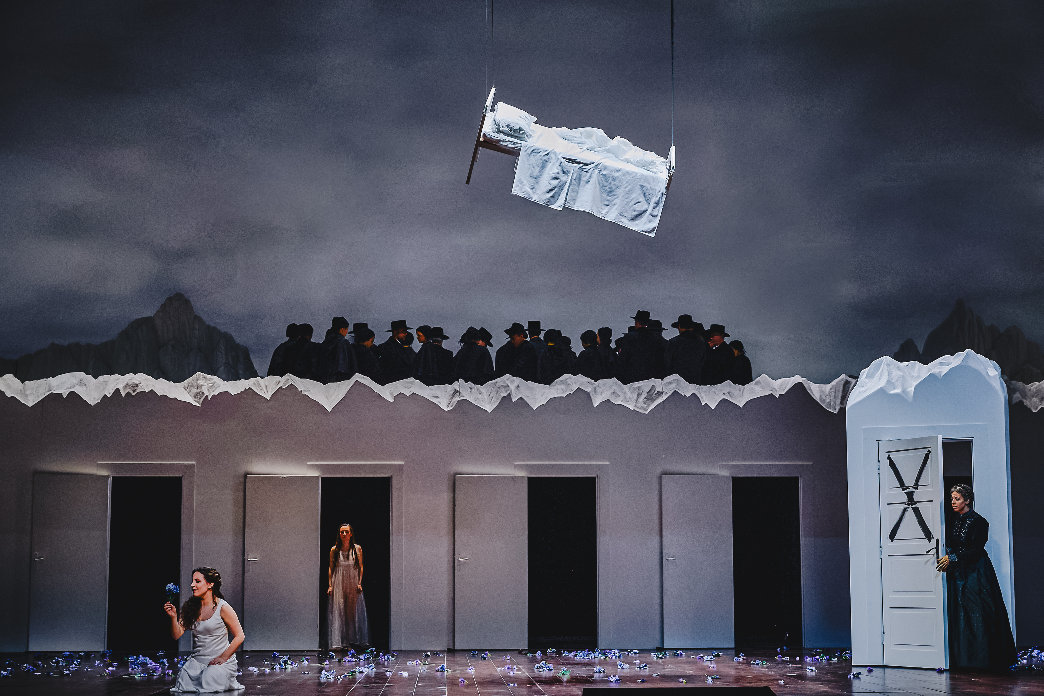 A surreal and theatrical stage scene from The Metropolitan Opera’s production of La Sonnambula. A bed with white sheets is suspended in mid-air, with a figure lying under the covers. Below, a group of people dressed in black cloaks and top hats stand in a row against a mountainous backdrop. The lower part of the stage features several open doors, from which women in flowing white dresses emerge. One woman kneels on the floor holding a blue flower, while another stands in a doorway. On the right side, a woman dressed in black peers out from behind a partially open door, which is marked with an ominous "X." Scattered blue flowers cover the wooden stage floor, adding to the eerie and dreamlike atmosphere.
