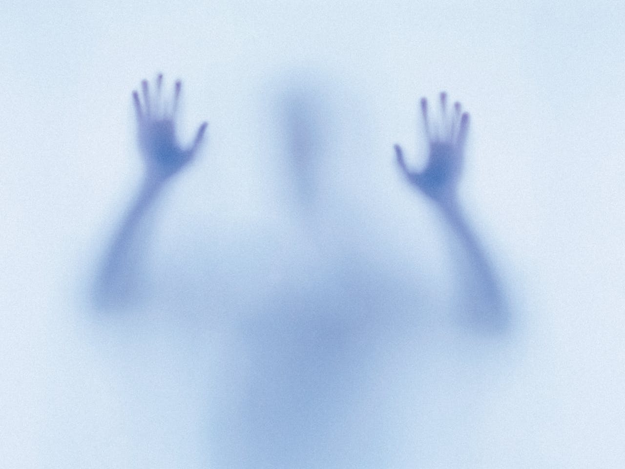 A mysterious and eerie image featuring the blurred silhouette of a person pressing their hands against a translucent surface. The figure appears to be reaching out or trapped, with their shadowy form creating a sense of tension and unease. The blue-toned lighting enhances the ghostly, dreamlike atmosphere, evoking themes of isolation or the supernatural. The image represents the children in Natalie Abrahami’s production of The Turn of the Screw.