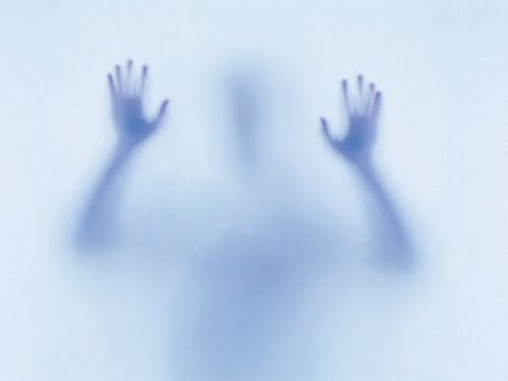 A mysterious and eerie image featuring the blurred silhouette of a person pressing their hands against a translucent surface. The figure appears to be reaching out or trapped, with their shadowy form creating a sense of tension and unease. The blue-toned lighting enhances the ghostly, dreamlike atmosphere, evoking themes of isolation or the supernatural. The image represents the children in Natalie Abrahami’s production of The Turn of the Screw.