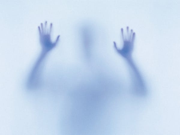 A mysterious and eerie image featuring the blurred silhouette of a person pressing their hands against a translucent surface. The figure appears to be reaching out or trapped, with their shadowy form creating a sense of tension and unease. The blue-toned lighting enhances the ghostly, dreamlike atmosphere, evoking themes of isolation or the supernatural. The image represents the children in Natalie Abrahami’s production of The Turn of the Screw.