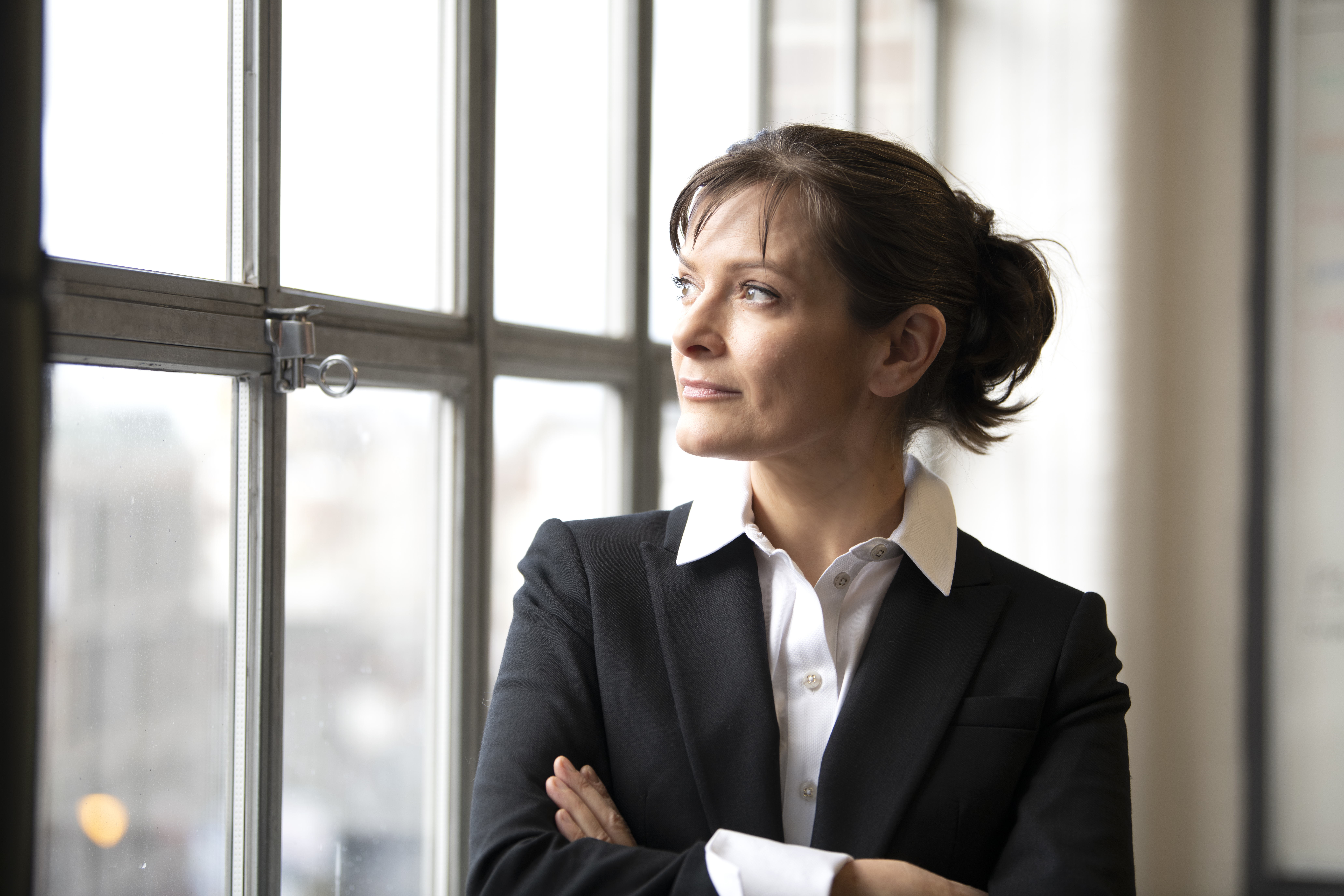 A person stands with their arms folded, looking out of a window. They have short brown hair that is tied up and they are wearing a white shirt and black blazer.