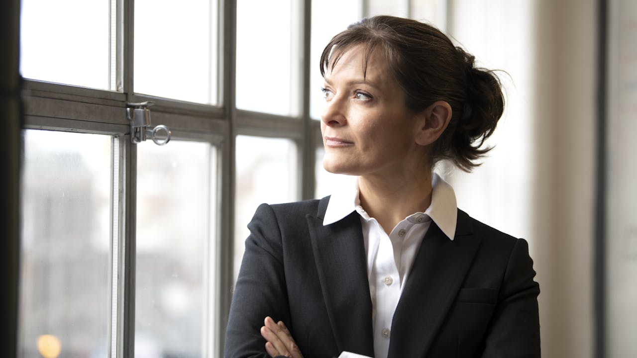 A person stands with their arms folded, looking out of a window. They have short brown hair that is tied up and they are wearing a white shirt and black blazer.