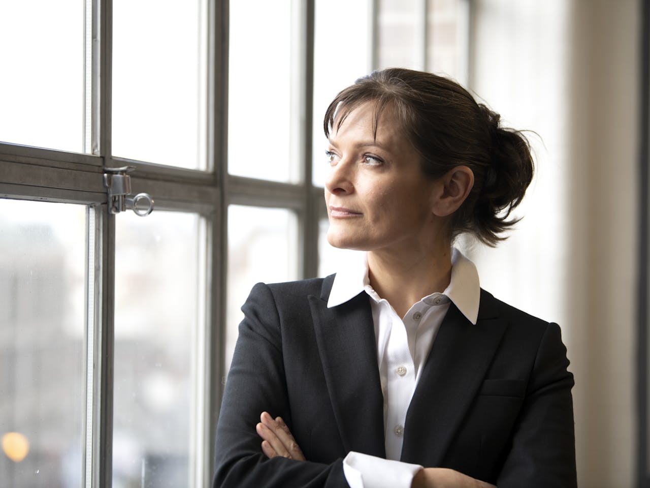 A woman stands with their arms folded, looking out of a window. They have short brown hair that is tied up and they are wearing a white shirt and black blazer.