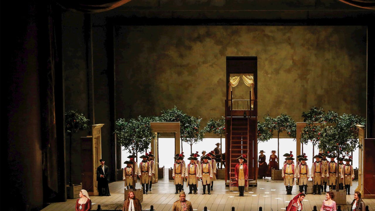 A view of the whole stage set for The Metropolitan Opera's production of THe Barber of Seville. Six performers stand at the front of the stage in 18th Century costume. At the back of the stage rows of soldiers are positioned. The stage is set with trees, open doorways and a staircase.