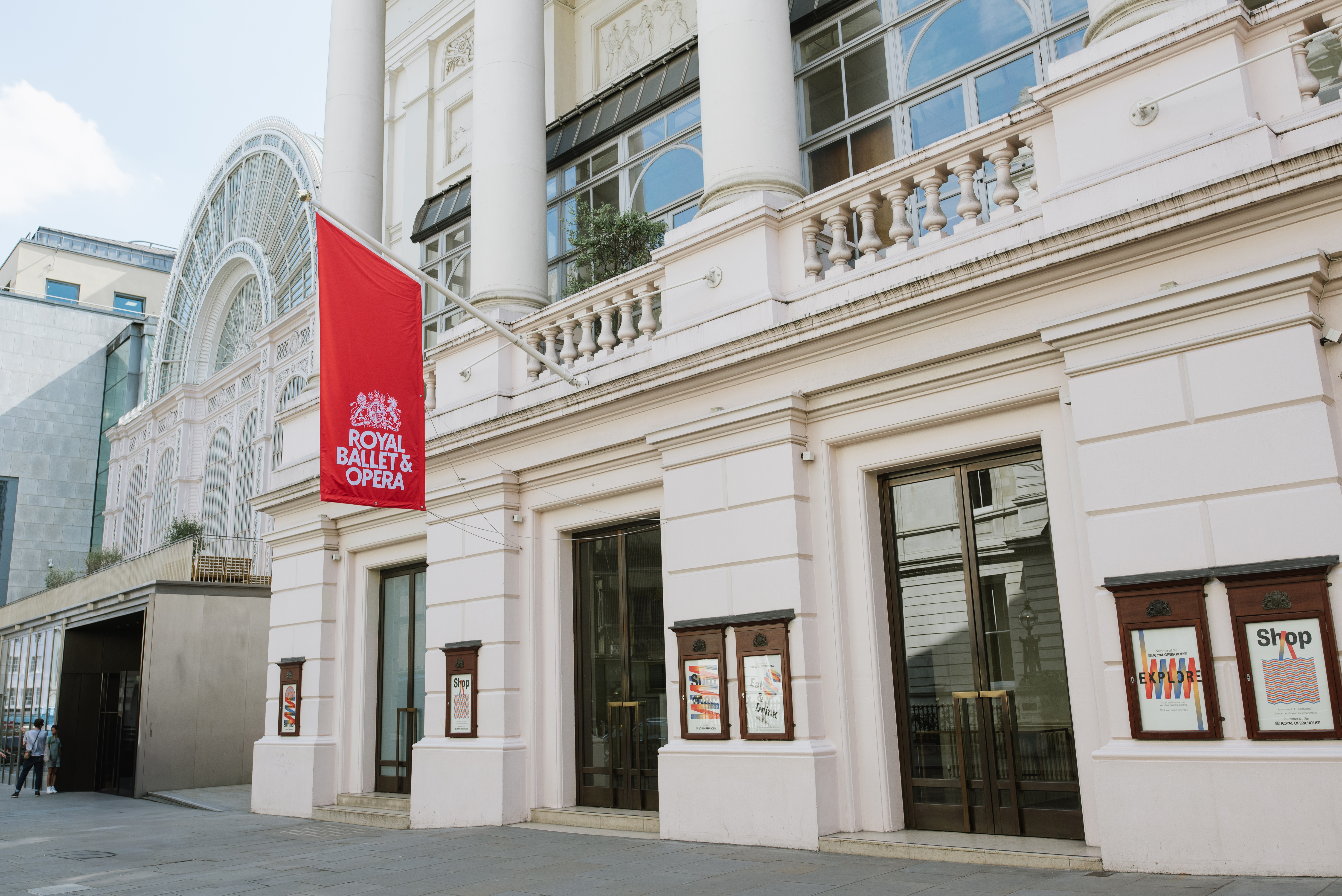 The facade of a Victorian building with columns and a red flat which sits next to a glass fronted building. It is the Royal Opera House in London
