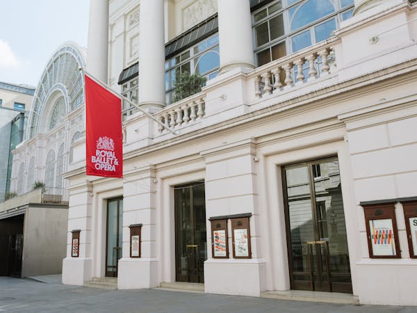 The facade of a Victorian building with columns and a red flat which sits next to a glass fronted building. It is the Royal Opera House in London