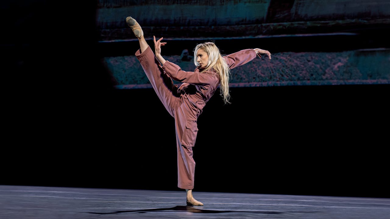 A woman with blonde hair and wearing a pink boiler suit lifts her leg above her head.