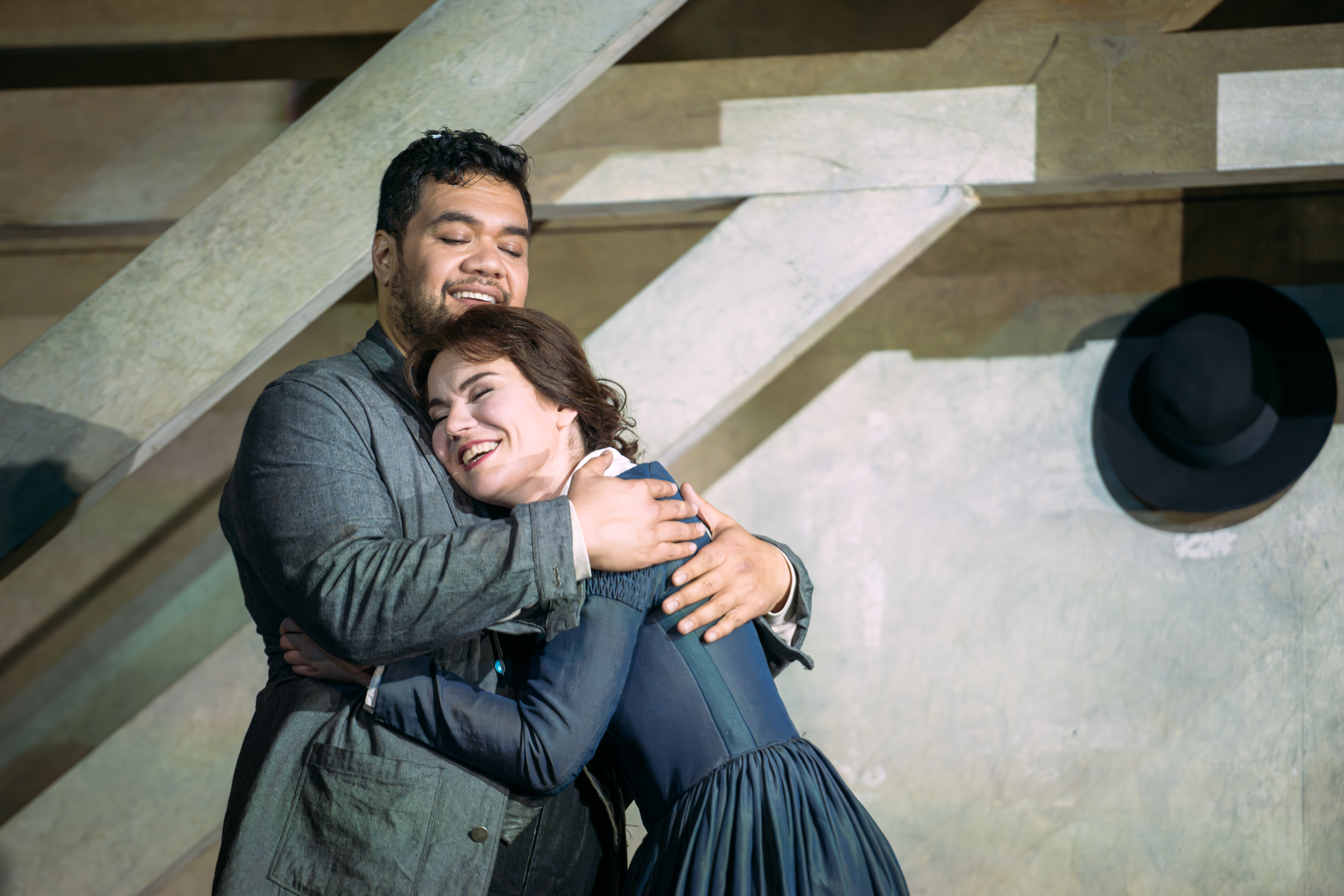 Two performers embrace on stage. They are in an attic room with wooden beams. The woman is wearing a blue dress and the man is wearing a grey jacket.