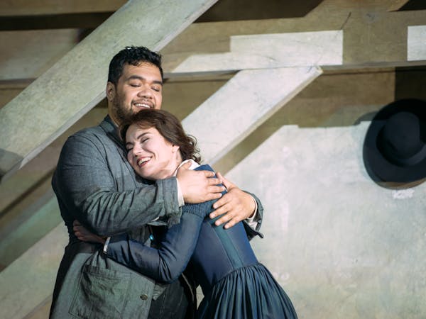 Two performers embrace on stage. They are in an attic room with wooden beams. The woman is wearing a blue dress and the man is wearing a grey jacket.