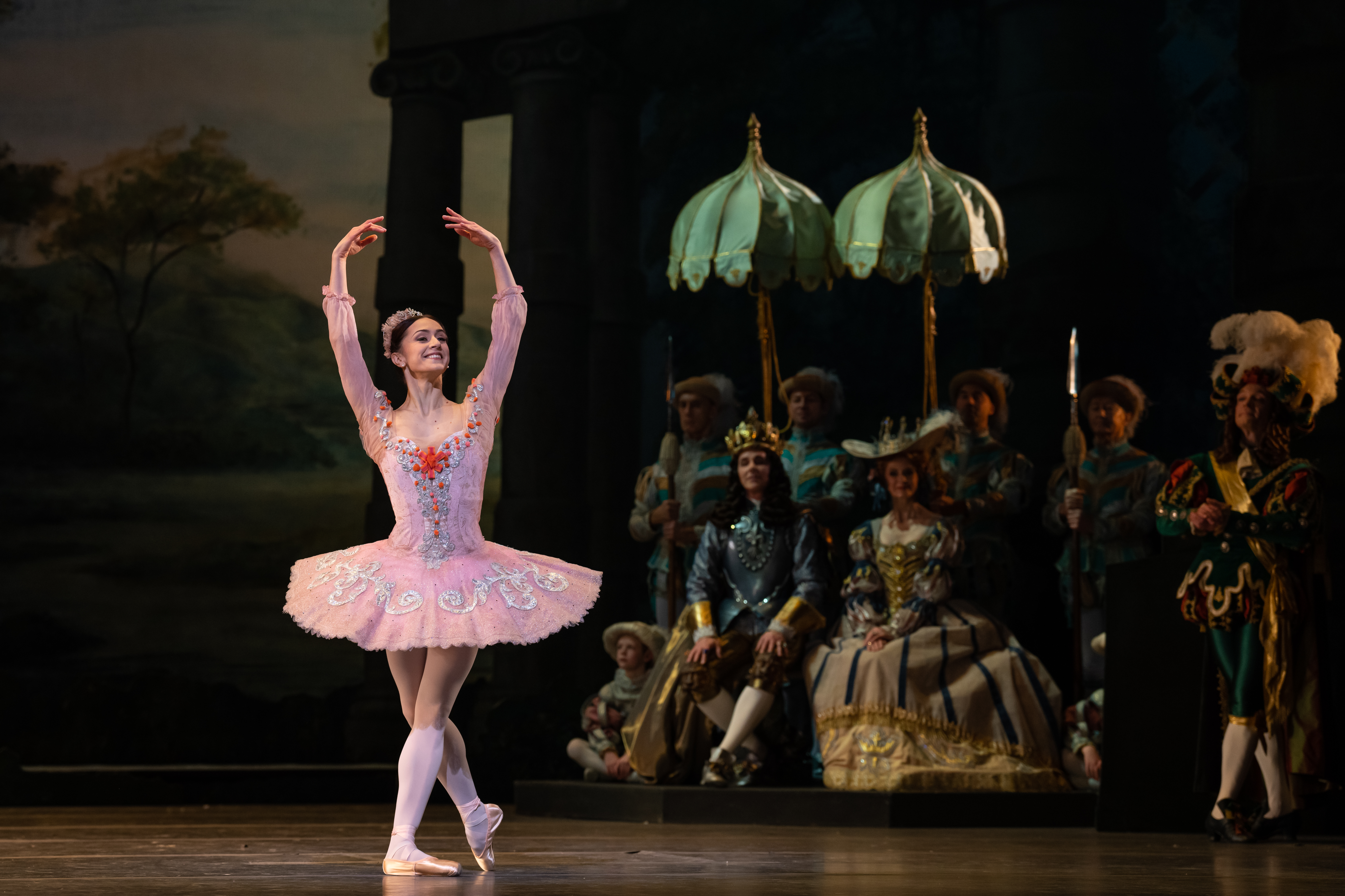Marianela Nuñez stands in the middle of the stage and holds her arms above her head in a pose. She is wearing a pink tutu and pink leotard. The tutu and leotard have silver and red detailing. 