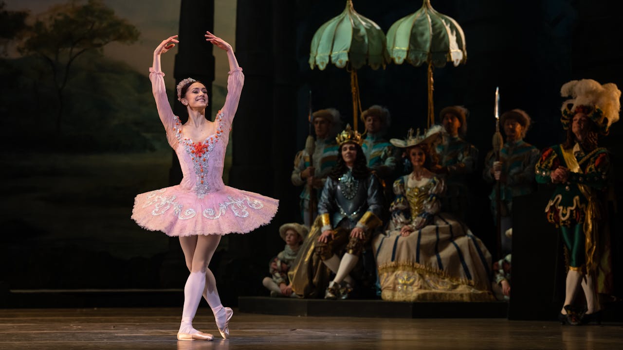 Marianela Nuñez stands in the middle of the stage and holds her arms above her head in a pose. She is wearing a pink tutu and pink leotard. The tutu and leotard have silver and red detailing.