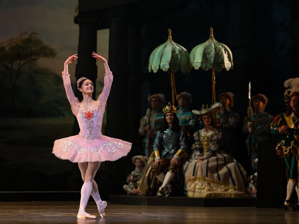 Marianela Nuñez stands in the middle of the stage and holds her arms above her head in a pose. She is wearing a pink tutu and pink leotard. The tutu and leotard have silver and red detailing.