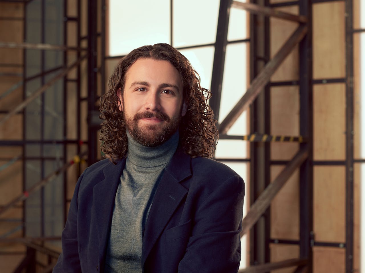 A man wearing a navy suit jacket over a grey turtleneck sits in a brightly lit studio with wooden panelled frame behind her. A headshot of Ryan Vaughan Davies.