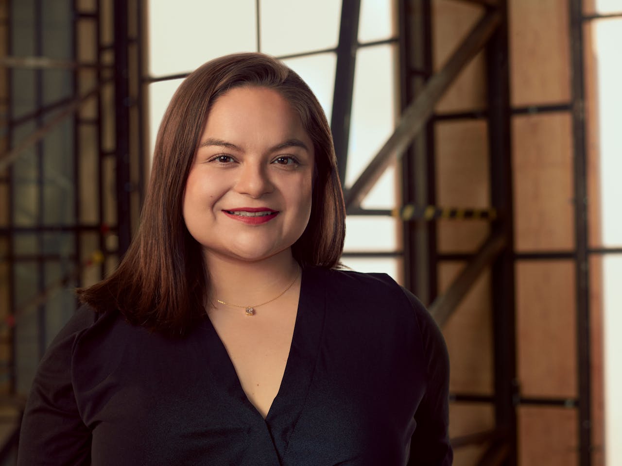 A woman wearing a dark shirt sits in a brightly lit studio with wooden panelled frame behind her. A headshot of Isabela Diaz.