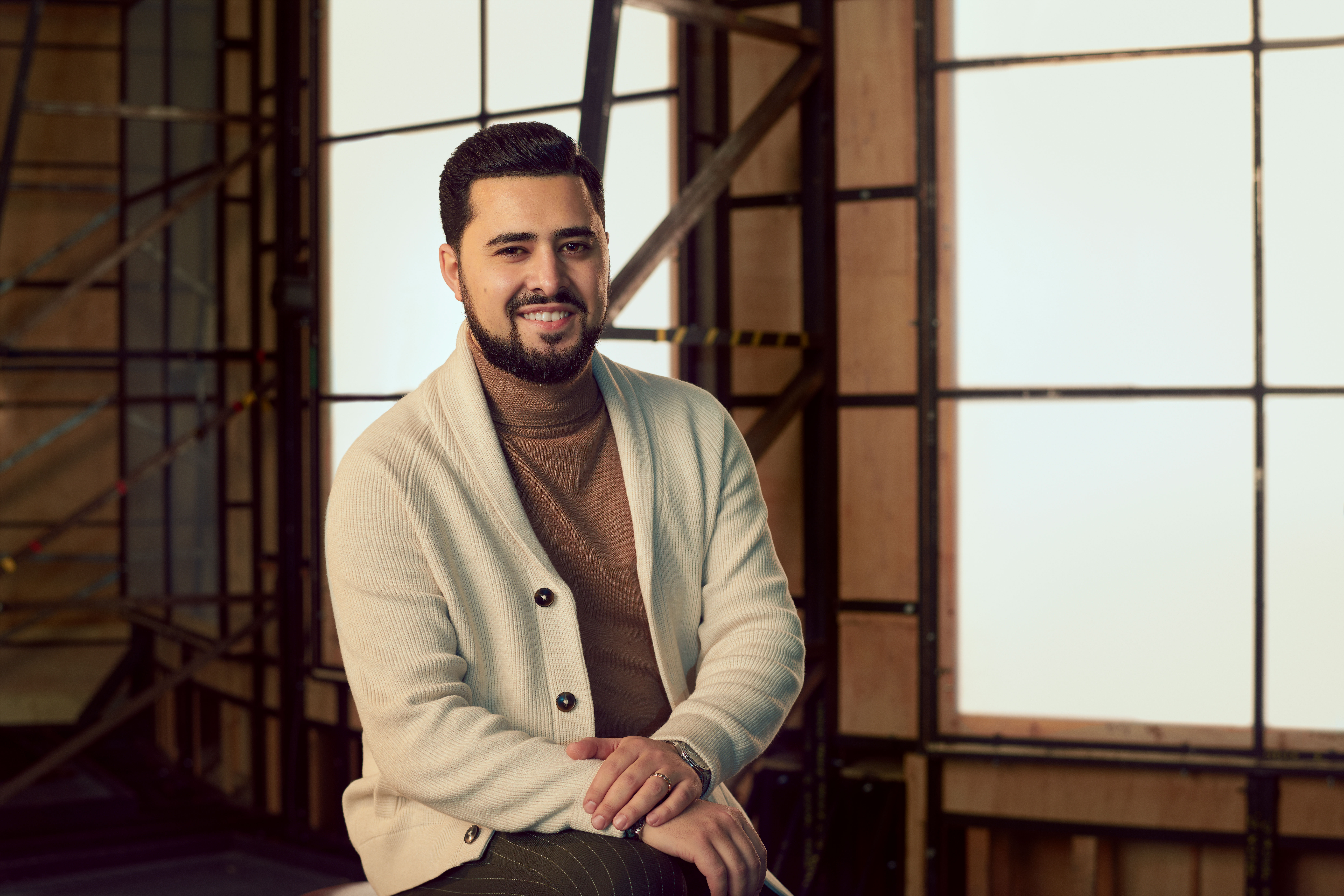 A man wearing a cream cardigan over a brown turtleneck, sits in a brightly lit studio with wooden panelled frame behind him. A headshot of Grisha Martirosyan. 