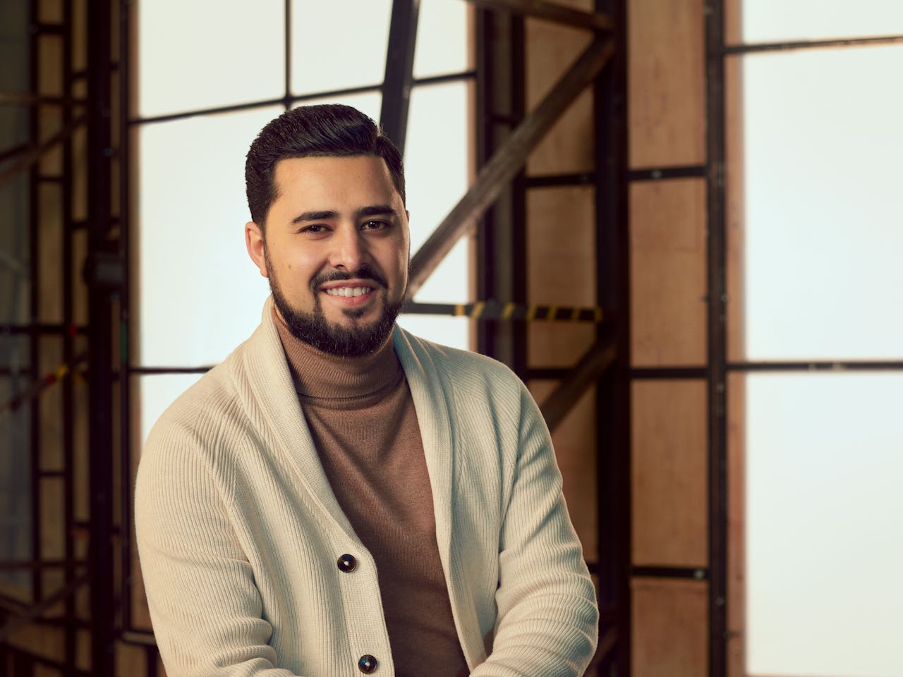 A man wearing a cream cardigan over a brown turtleneck, sits in a brightly lit studio with wooden panelled frame behind him. A headshot of Grisha Martirosyan.