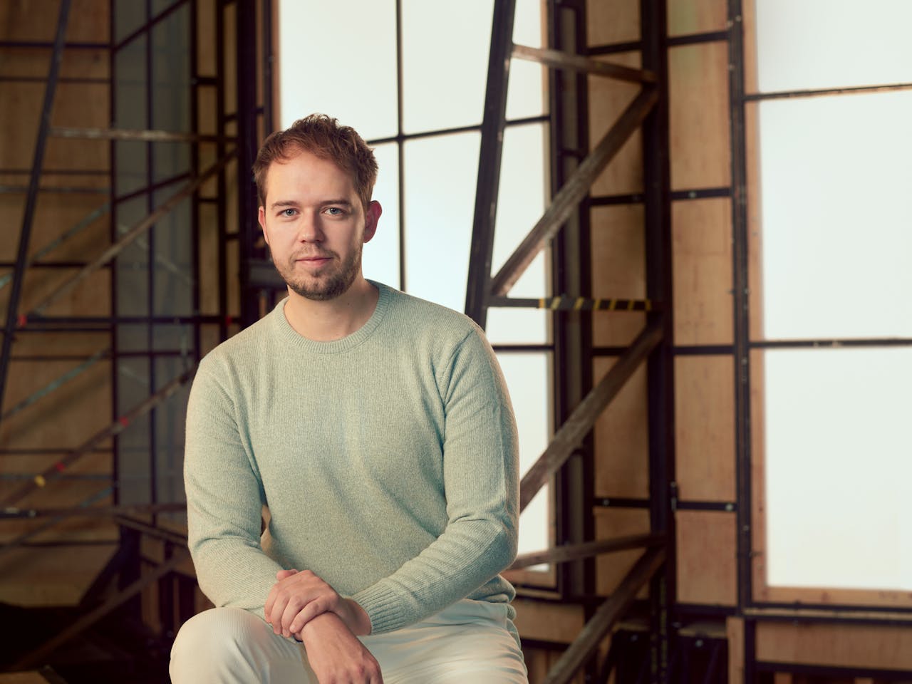 A man wearing a pale green jumper sits in a brightly lit studio with wooden panelled frame behind her. A headshot of Jamie Woollard.