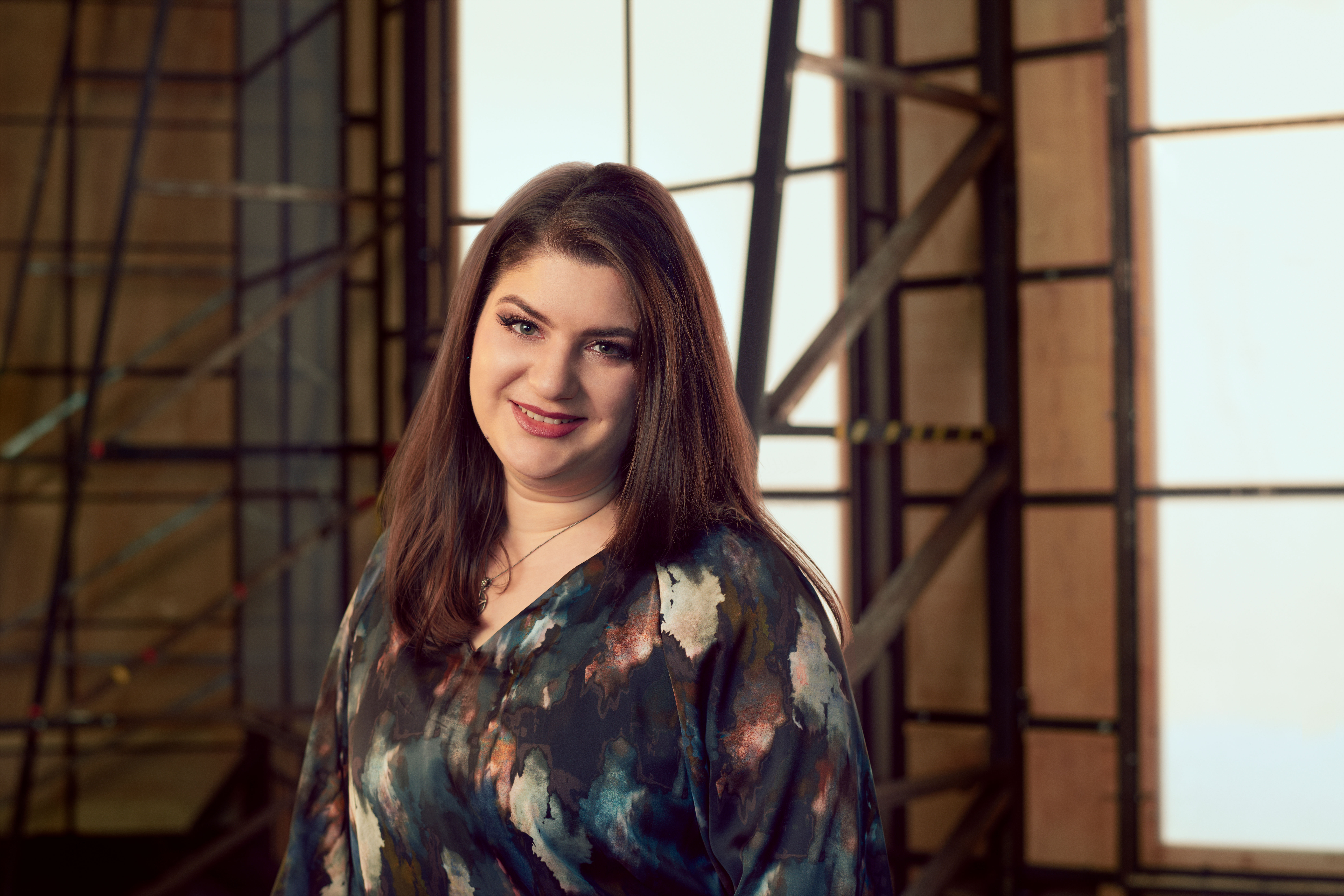 A woman wearing a dark patterned shirt sits in a brightly lit studio with wooden panelled frame behind her. A headshot of Valentina Puskas. 