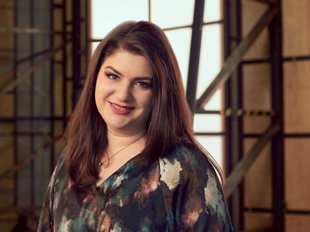 A woman wearing a dark patterned shirt sits in a brightly lit studio with wooden panelled frame behind her. A headshot of Valentina Puskas.