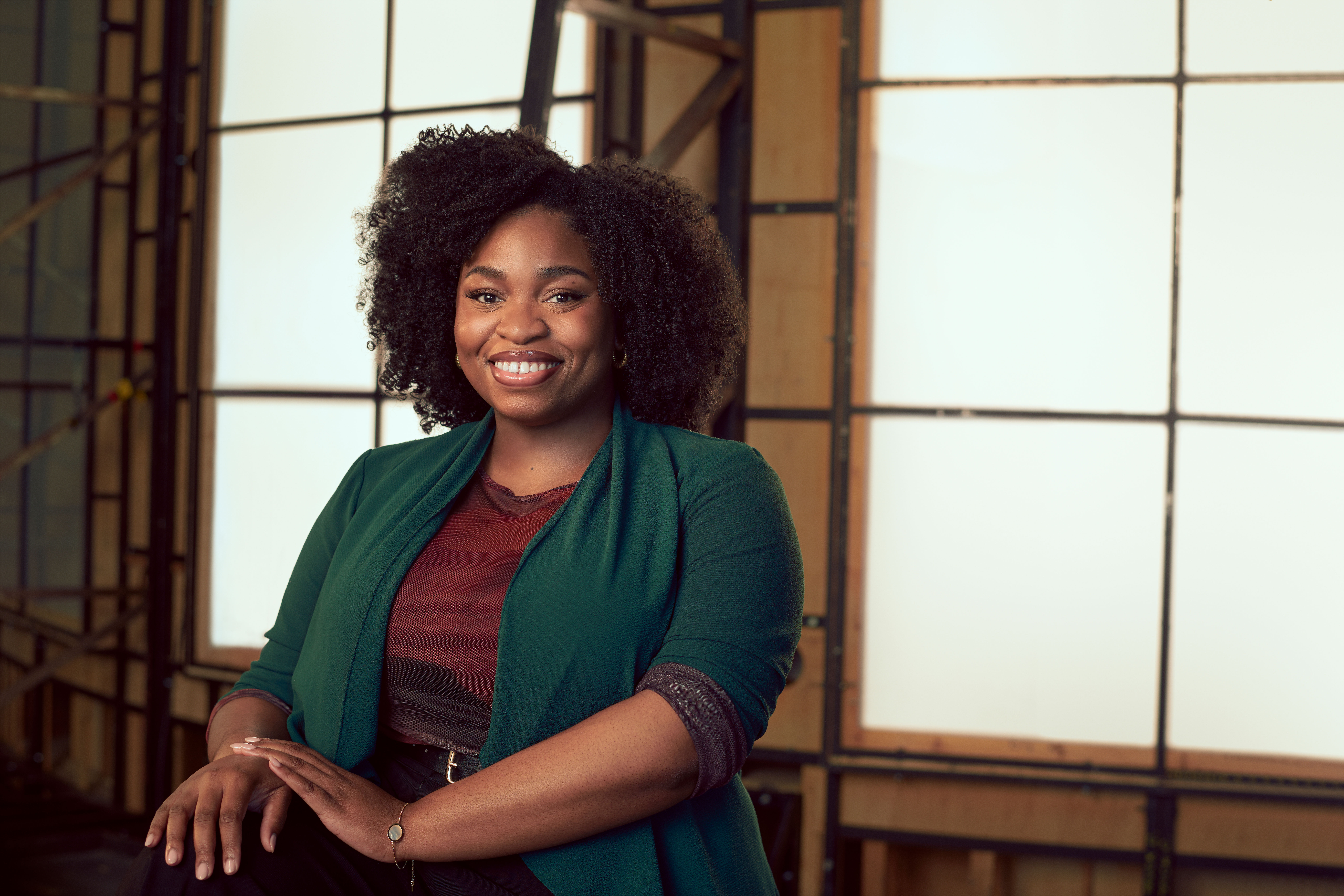 A woman wearing a teal cardigan over a brick red shirt sits in a brightly lit studio with wooden panelled frame behind her. A headshot of Veena Akama Makia.  