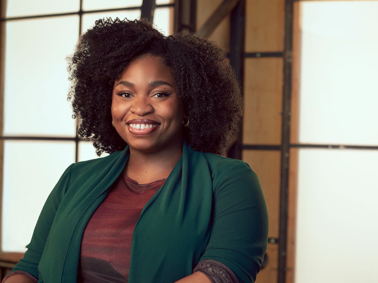 A woman wearing a teal cardigan over a brick red shirt sits in a brightly lit studio with wooden panelled frame behind her. A headshot of Veena Akama Makia.