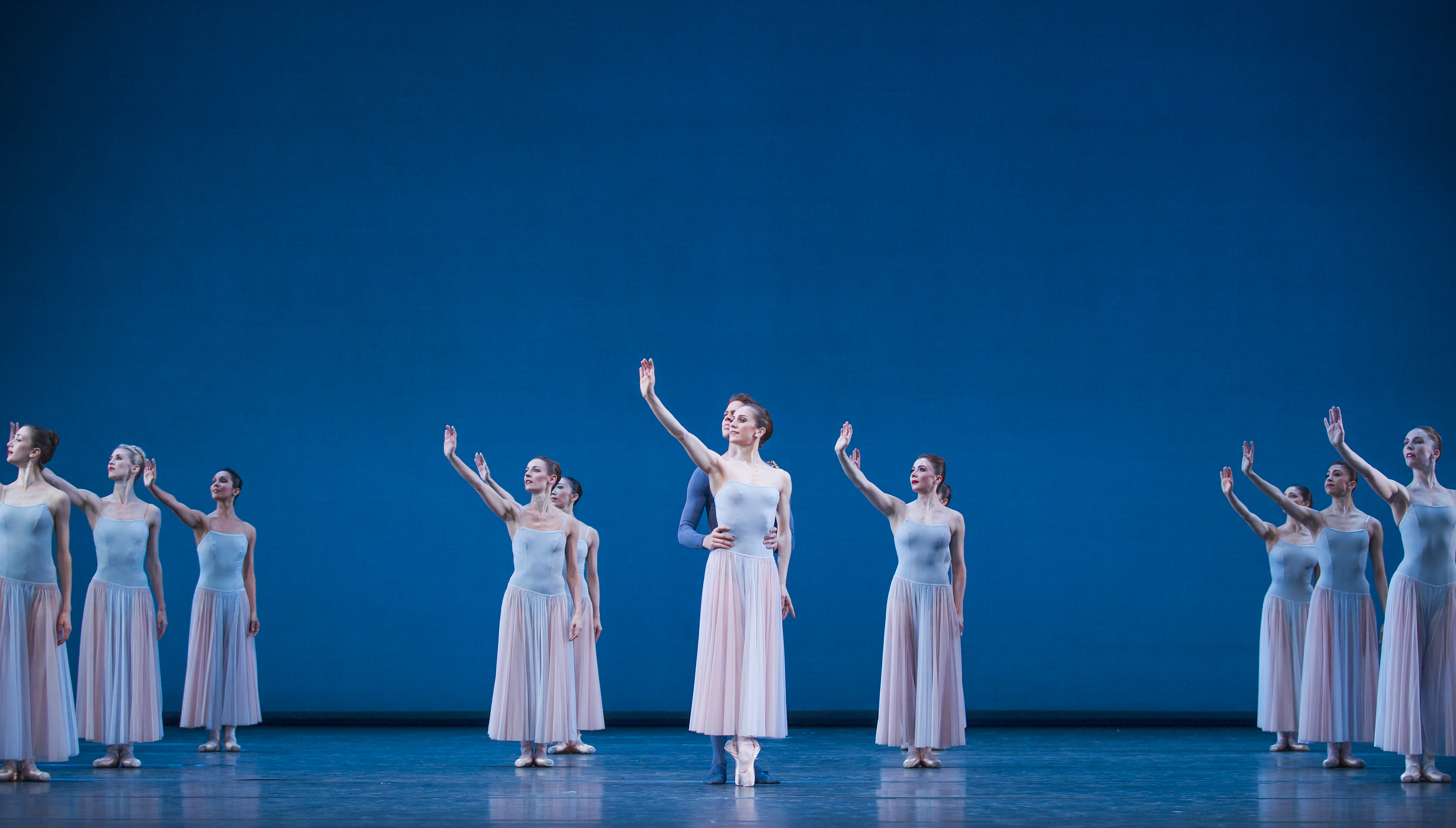10 ballet dancers stand on a blue-set stage holding their right arms in the air. In the centre, there are 5 dancers and either side of hte stage are three dancers - all are wearing skirts. The dancer at the front is being held by the waist by another dancer wearing blue trousers and top.

