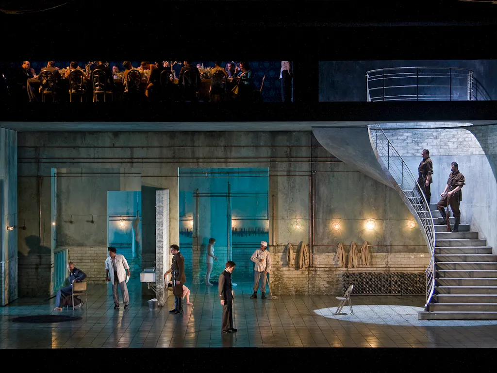 A modern stage set features actors in various poses on a realistic, industrial-style set with stairs and blue lighting; an orchestra is visible above the main stage area.