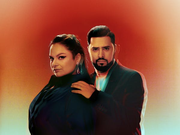 A person wearing all black stands sideways and another person who is also wearing all black holds their shoulder with their left hand facing the front. The background is set with gradient colours starting with orange moving down to blue-green.