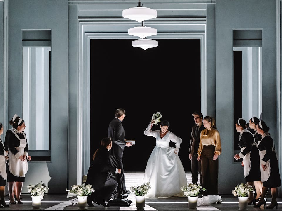 The stage is set with a large modernist concrete doorway with a tall window either side, three circular lights on the ceiling and five white flowerpots filled with white flowers. In the centre of the doorway, a performer dressed as a bride in a white dress holds up a bouquet of flowers in an angry pose, her veil sits on the floor. A chorus of performers, some of them are dressed in black and white maid uniforms and others in smart suits, look on.