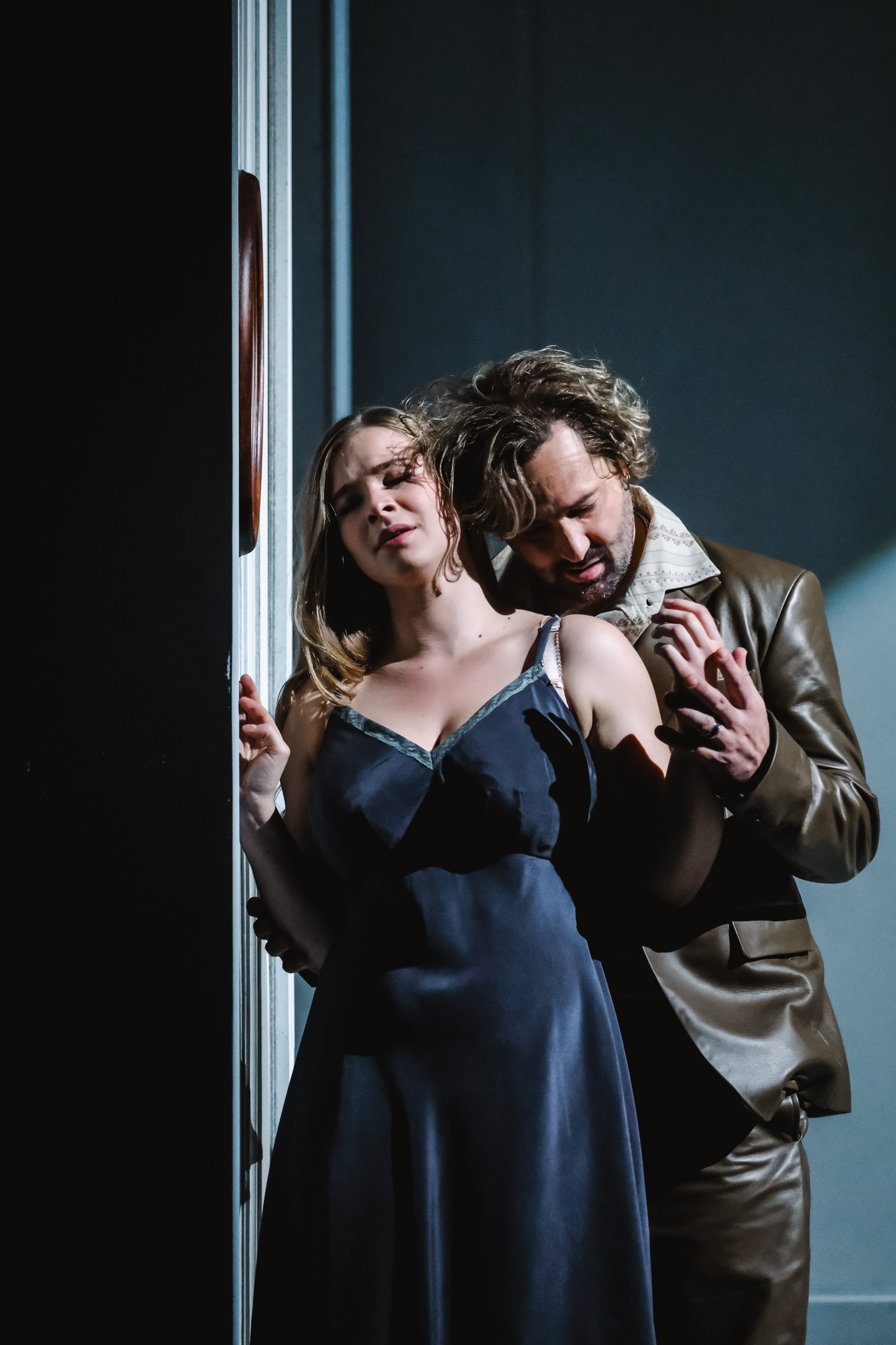 A performer wearing a navy-blue silk dress stands near a wall. Their eyes are closed, and their hands are being stroked from behind in a passionate embrace by another performer wearing a brown leather jacket and trousers and patterned beige shirt. His head almost rests gently on her shoulder. 